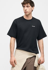 Νέος άνδρας που φοράει μαύρο t-shirt Columbia, μπεζ μπουφάν δεμένο γύρω από τη μέση, κρατώντας ένα ταιριαστό μπεζ καπέλο, κοιτάζοντας πλάγια σε λευκό φόντο.