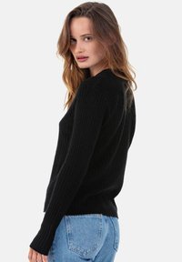 Pull en maille côtelée noir avec manches longues et coupe ajustée, associé à un jean bleu clair. La texture est visible avec un motif subtil.