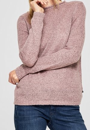 Žena v světle fialovém pleteném svetru s dlouhými rukávy a modrých džínách, pózující s jednou rukou u obličeje.