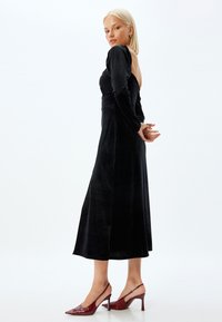 Mustapellavillainen pitkä mekko pitkillä hihoilla ja avonaisella selällä, yhdistettynä burgundin punaisiin teräskärjellisiin slingback-korkokenkiin. Silkkinen rakenne ja virtaava muotoilu.