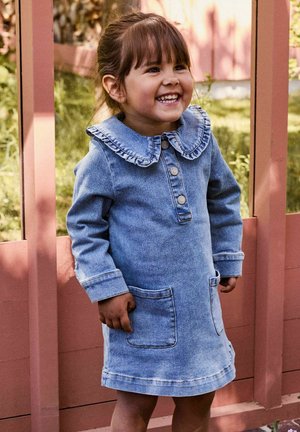 Bambino sorridente che indossa un vestito di jeans blu con un colletto arricciato, in piedi all'aperto davanti a una struttura in legno rosa.