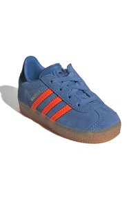Blå fløyelsneaker med oransje tre striper, rund tå og teksturert gummisåle. Svart hælforsterkning til stede.