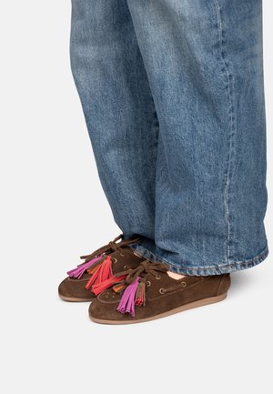 Zapatos mocasines de ante marrón con borlas moradas y rojas, usados con vaqueros anchos azules sobre un fondo liso.
