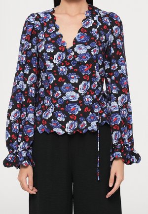 Femme portant un chemisier noir cache-cœur à motif floral bleu, blanc et rouge, col en V festonné, manches bouffantes et poignets à volants.