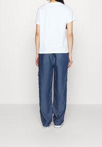 Mujer de pie, de espaldas, vistiendo una camiseta blanca de manga corta sencilla, pantalones cargo azules holgados con bolsillo lateral y zapatillas blancas.