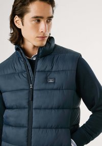 Marineblå puffervest med høy krage, glidelås, og horisontal søm; merket label på forsiden; brukt over en mørk genser.