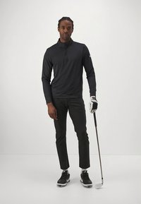 Fekete, hosszú ujjú póló fél cipzárral, hozzáillő, fekete, testhezálló nadrággal párosítva. A személy egy golfütőt tart, fekete sportcipőt és golfkesztyűt visel.