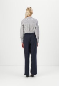 Femme aux longs cheveux blonds, debout face à un mur blanc, portant une chemise rayée à manches longues rentrée dans un pantalon noir à jambes larges et des talons noirs.