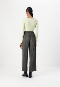 Donna con capelli intrecciati che indossa un maglione a coste verde pallido, pantaloni grigi a pieghe a vita alta e stivali neri con tacco, di spalle.