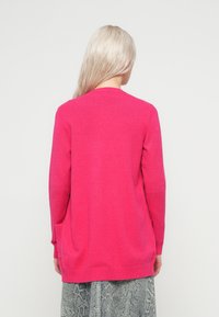 Cardigan lungo fucsia a maniche lunghe realizzato in tessuto morbido, con orlo a coste e design aperto. Il modello indossa una gonna grigia dallo stile testurizzato.