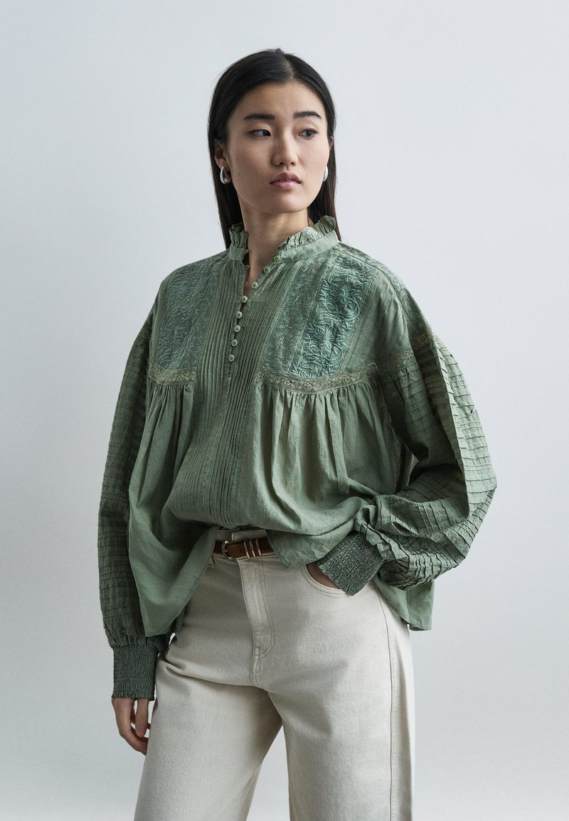 Νεαρή γυναίκα με ίσια σκουρόχρωμα μαλλιά, που φοράει μια φαρδιά πράσινη κεντητή μπλούζα και εκρού παντελόνι με καφέ ζώνη, στέκεται μπροστά σε απλό φόντο.