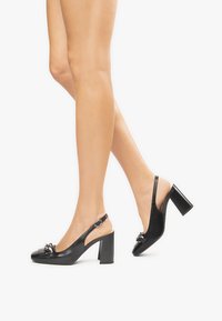 Dei sandali slingback neri in pelle con punta quadrata, dotati di un tacco spesso, un dettaglio a fiocco e una cinghia regolabile dietro la caviglia.