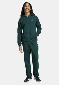 Felpa con zip verde scuro e pantaloni da jogging abbinati in materiale morbido, con il logo Adidas in una tonalità a contrasto sul petto e sulla gamba.