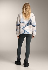 Femeie văzută din spate, purtând o jachetă texturată albă și albastră cu un design de peisaj de iarnă, colanți închiși la culoare, șosete gri și cizme negre de iarnă.