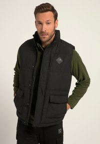 Schwarze gepolsterte Weste mit hohem Kragen, Fronttaschen und einem Diamantlogo auf der Brust, getragen über einem grünen Langarmshirt.