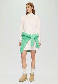 Cremefarbendes Pulloverkleid mit gerader Schnittführung; grünes Strickwickel mit Knoten in der Taille; beige Stiefeletten; hellgraue Socken. Einfaches und lässiges Design.