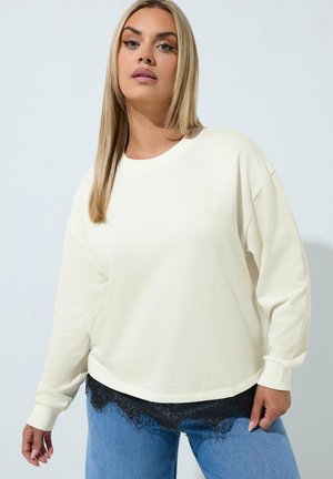 Kobieta o długich blond włosach, ubrana w biały top z długim rękawem, ozdobiony czarną koronką, oraz niebieskie dżinsy, stojąca na jednolitym jasnym tle.