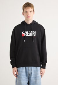 Svart hettegenser med frontlomme, med en flerfarget "CALVIN KLEIN" logo. Laget av myk stoff med en avslappet passform.