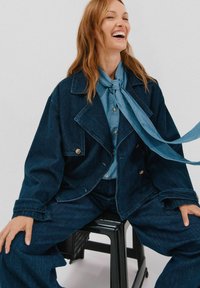 Kurtka dżinsowa z wyraźnym kołnierzem, niebieskimi guzikami i kieszeniami na łatki, w połączeniu z niebieską koszulą na guziki i pasującą krawatem.