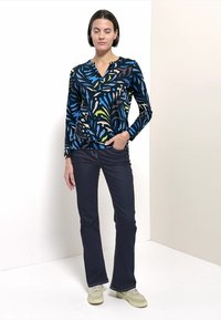 Langärmeliges Oberteil mit dunklem Hintergrund und bunten Blattmustern, kombiniert mit dunkler Jeans und hellen Sneakern. Glatte Stoffstruktur.