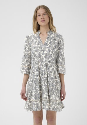 Jeune femme aux cheveux clairs portant une robe blanche longueur genou avec imprimé floral noir et manches trois-quarts avec un col en V.