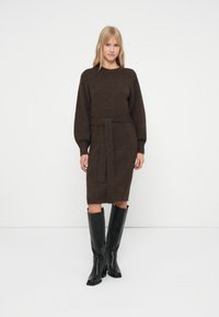 Donna che indossa un abito lavorato a maglia marrone scuro con cintura, maniche lunghe e stivali di pelle neri fino al ginocchio, su uno sfondo bianco semplice.