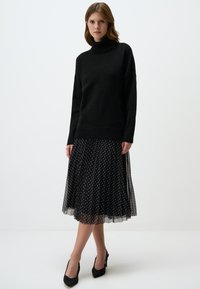 Maglione nero a collo alto con orlo a costine, abbinato a una gonna nera a pois plissettata e scarpe con tacco alto nere.