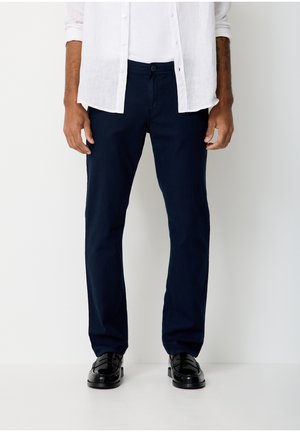 Man wearing navy blue pants, black leather shoes, white shirt, and unbuttoned white long-sleeve shirt, standing against a plain background.