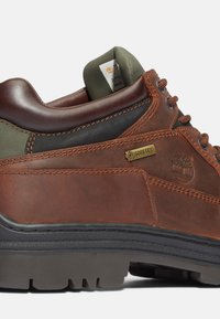 Brūni ādas pārgājienu apavi ar zaļiem akcentiem, teksturēta virsma, Gore-Tex zīmols, kontrastējoša šuvju krāsa un melna gumijas zole.