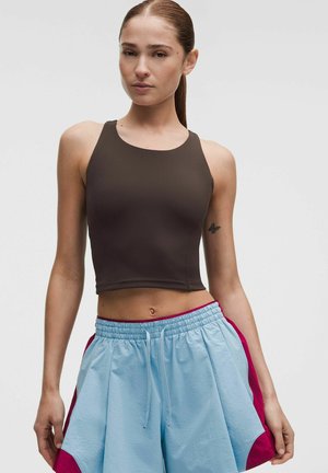 Junge Frau mit braunem ärmellosem Crop-Top und hellblauen Sportshorts mit roten Akzenten, stehend vor einem einfarbigen Hintergrund.