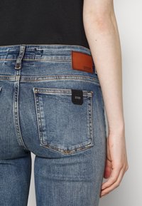 Jeans de denim azul con un lavado ligeramente descolorido, que presentan un parche de cuero marrón en la parte posterior y una etiqueta de marca negra cerca del bolsillo.