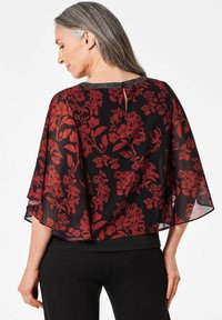 Top nero trasparente con motivo floreale rosso, maniche a batwing fluide e una sottile apertura a chiave sul retro. Abbinato a pantaloni neri aderenti.