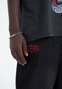 Černé bavlněné tričko s barevným designem z kamínků, v kombinaci s tmavými džínovými kalhotami s červenou vyšívanou značkou a stříbrným náramkem s řetízkem.