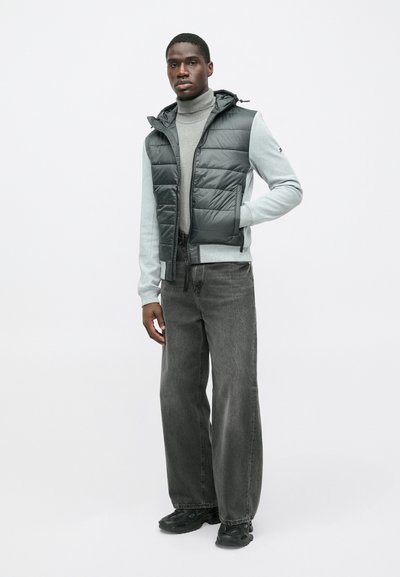 Pilkas pūkuotas liemenė su gobtuvu, vilkima virš šviesiai pilko golfo, derinama su laisva tamsiomis džinsomis ir juodais sportbačiais, su užtrauktuku priekyje.