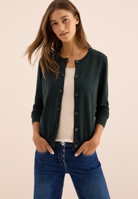 Frau mit langen braunen Haaren, die einen dunkelgrünen, knöpfbaren Cardigan über einem beigen Top und blauen Jeans trägt, mit Händen in den Taschen auf beigem Hintergrund.