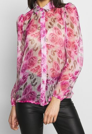 Femme portant un chemisier transparent rose à fleurs boutonné, avec des épaules bouffantes et des manches longues, associé à un pantalon en cuir noir.