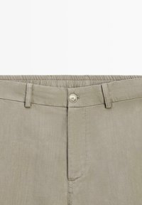 Beżowa talia spodni z zapięciem na guzik, szlufkami na pasek i częściowo elastycznym pasem na jednolitym tle.