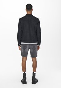 Svart mockajacka med krage, bärs över en randig skjorta, grå denimshorts och svarta ankelskor. Inga synliga mönster eller hårdvarudetaljer.