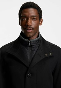 Uomo con capelli ricci corti e baffi, indossante un cappotto nero sovrapposto a una giacca trapuntata con collo alto, che guarda dritto contro uno sfondo semplice.