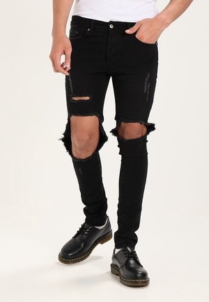 Muž v černých roztrhaných úzkých džínách s velkými dírami na kolenou a černých kožených botách na šněrování, stojící s jednou rukou v kapse.