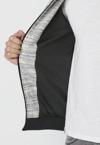 Giacca nera con zip, foderata in rete interna, con dettagli color grigio chiaro melange e orlo a coste neri. È visibile una mano che afferra la cerniera.