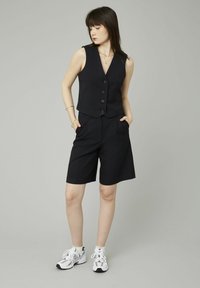 Jeune femme en gilet sans manches noir à rayures, shorts assortis jusqu'aux genoux, mains dans les poches, baskets blanches, regardant vers le bas à gauche.