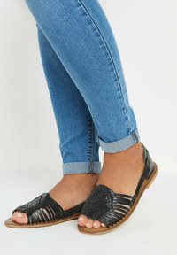 Mustat kudotut sandaalit, joissa on avonäinen kärki, litteapohja ja teksturoitu pinta, yhdistettyinä rullatuiksi käännettyihin sinisiin farkkuihin.