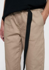 Beige katoenen broek met een relaxed fit, voorzien van zakken aan de voorkant, een zwart stoffen riem en een subtiel geborduurd nummer "10" op het been.