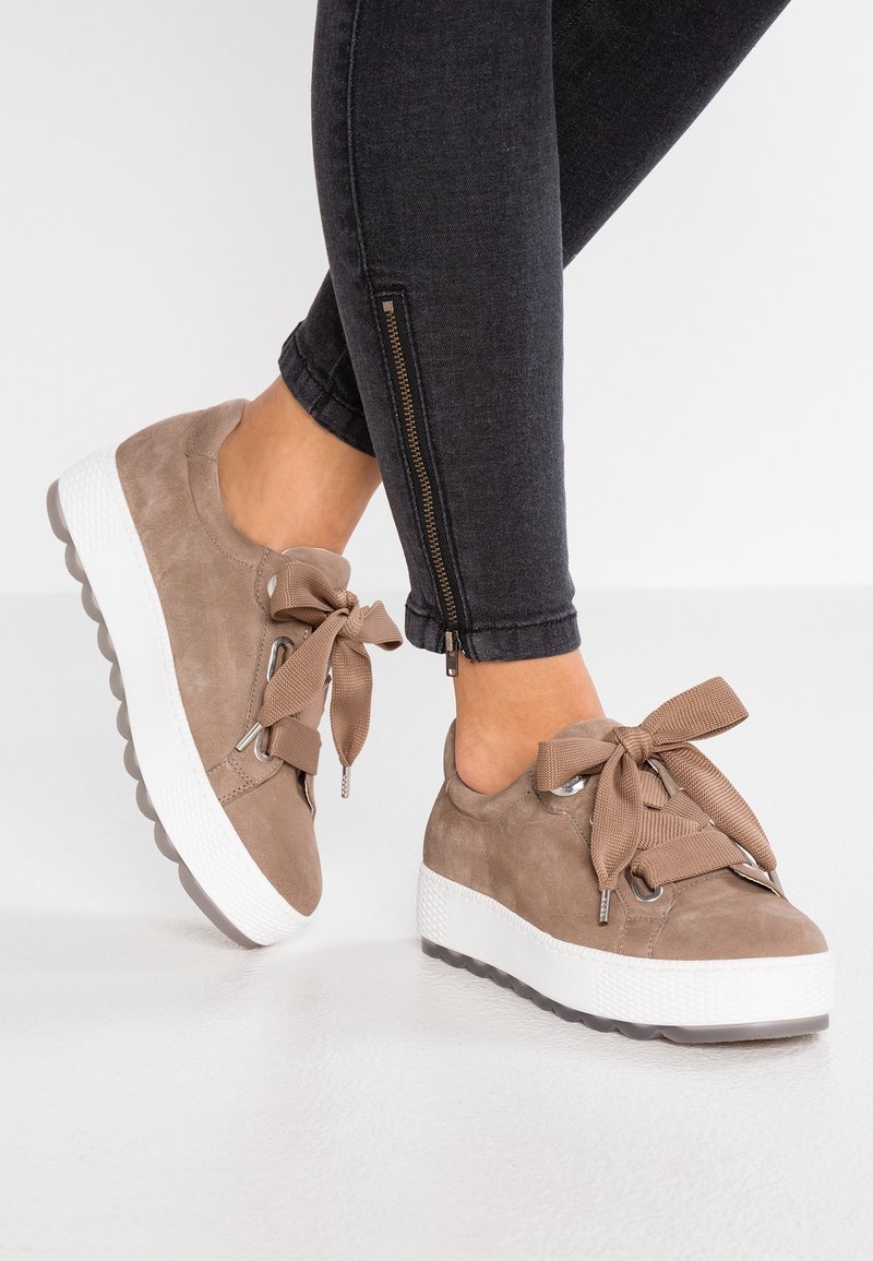 Personne portant des baskets compensées en daim taupe avec de larges lacets et un jean skinny noir avec des fermetures éclair latérales sur un sol blanc.