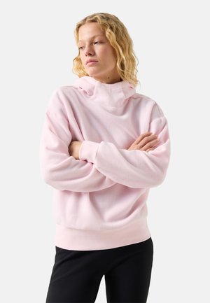 Žena s kudrnatými blond vlasy, oblečená v světle růžové mikině s kapucí a černých kalhotách, s překříženýma rukama, která se dívá stranou.