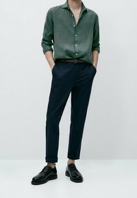 Camicia verde scuro con bottoni in tessuto strutturato, abbinata a pantaloni blu navy e scarpe nere. I pantaloni sono leggermente risvoltati.