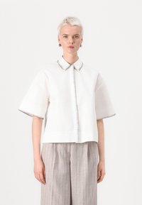 Camicia bianca a maniche corte con colletto a punta, caratterizzata da cuciture di contrasto nere lungo il colletto e l'orlo. Tessuto di cotone liscio.