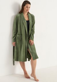 Vihreä fleece-haalari pitkillä hihoilla, kauluksella ja sivutaskuilla; sisältää vyön ja polvipituisen leikkauksen pehmeällä koostumuksella.