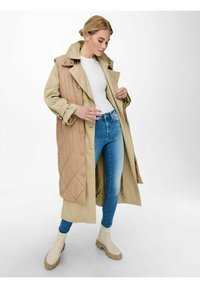 Beige quiltet kåpe lagt over en lys beige trenchcoat, kombinert med blå denim jeans og kremfargede ankelboots. Rene linjer og myke teksturer.